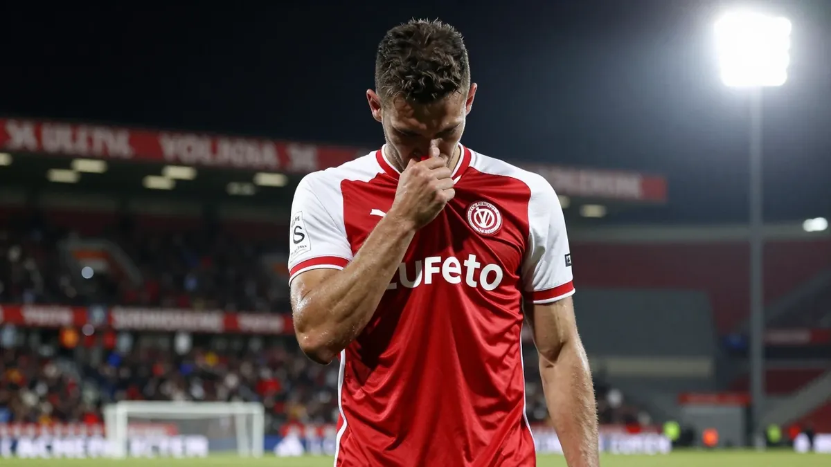A 25-year-old FC Volendam player, with a muscular build, wearing a red and white jersey, looking dejected after missing a...