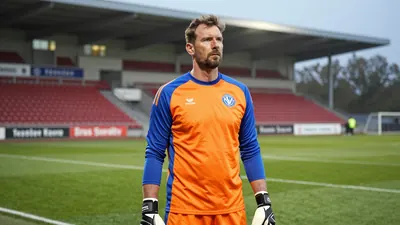 A tight, waist-up shot of a mid-30s, ruggedly-built goalkeeper with a weathered face, dressed in a plain orange and blue...