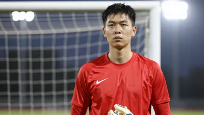 A 28-year-old goalkeeper in a red jersey with long sleeves, standing in a goal with a determined expression, wearing gloves