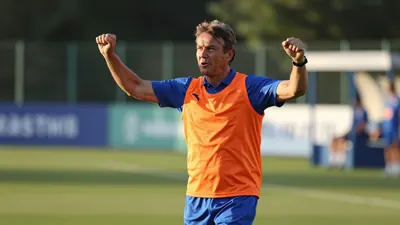 A 45-year-old coach in an orange and blue training top with a blue collar, mid-celebratory stride, arms raised, standing...